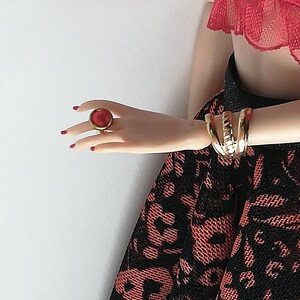 Op de afbeelding: Een rode ring met een gouden zetting op een vinger. De ring is op een hand die een zwarte en rode kanten rok en een rood kant topje draagt.