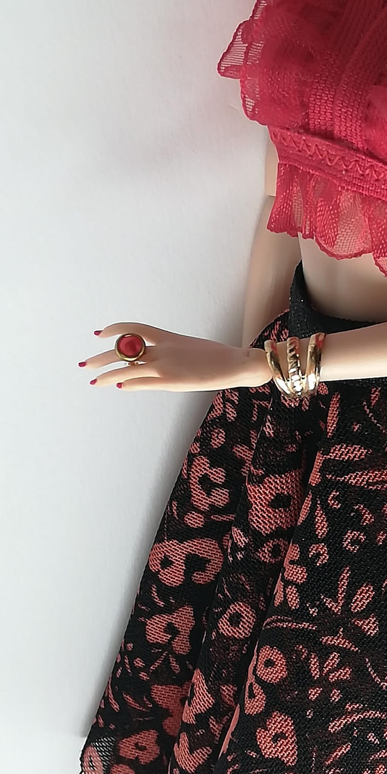 Op de afbeelding: Een rode ring met een gouden zetting op een vinger. De ring is op een hand die een zwarte en rode kanten rok en een rood kant topje draagt.