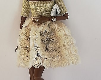 May include: A fashion doll with dark brown skin and long black hair styled in a braid. She is wearing a gold and white dress with a floral design and matching gold and white shoes.