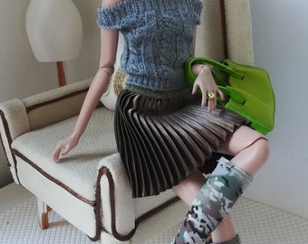 May include: A fashion doll wearing a grey sweater, a brown pleated skirt, and camouflage boots. The doll is sitting on a white armchair with a green handbag in her hand.
