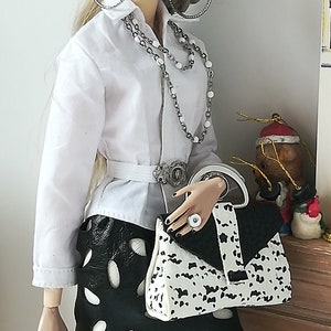 May include: A fashion doll wearing a white button-down shirt, a black skirt with white polka dots, and a white and black handbag. The doll has blonde hair and is wearing silver hoop earrings and a silver necklace.