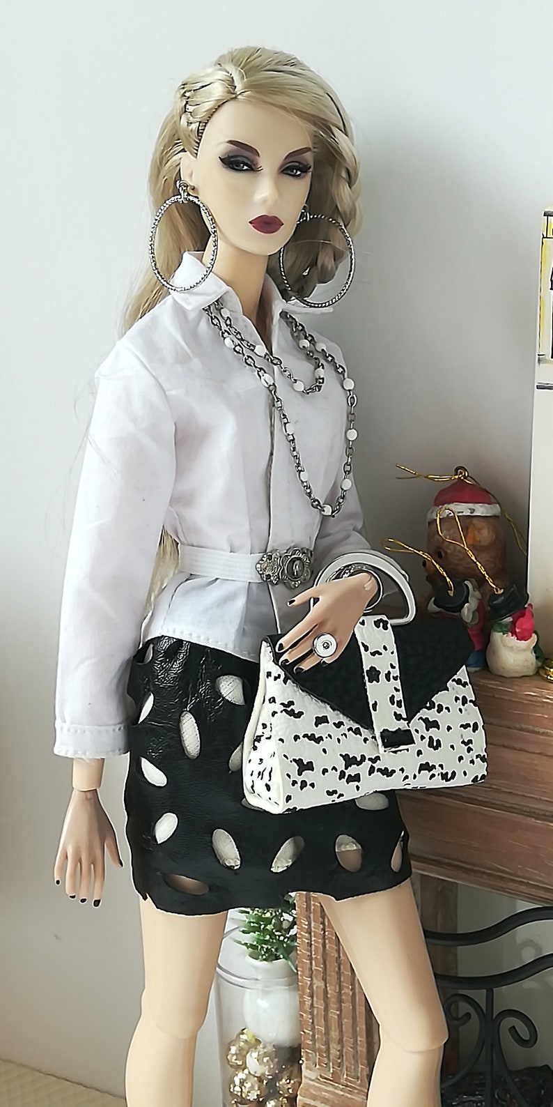 May include: A fashion doll wearing a white button-down shirt, a black skirt with white polka dots, and a white and black handbag. The doll has blonde hair and is wearing silver hoop earrings and a silver necklace.