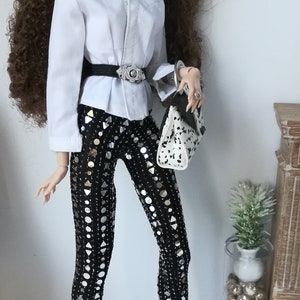 May include: A fashion doll with long brown curly hair wearing a white button-down shirt, a black belt with a silver buckle, black pants with silver sequins, black boots, and sunglasses. The doll is holding a white and black handbag.