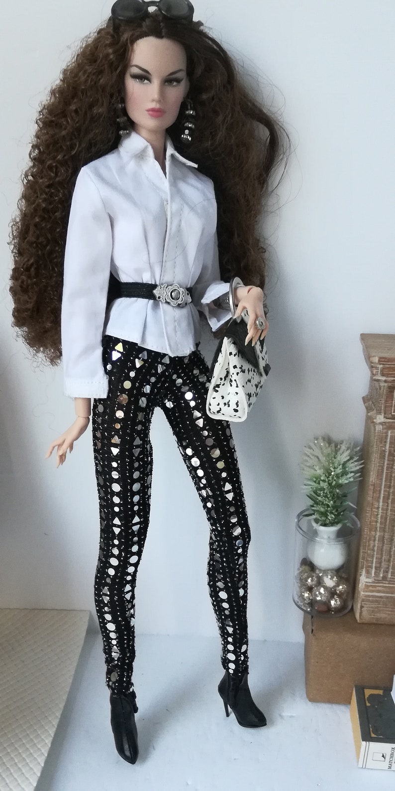 May include: A fashion doll with long brown curly hair wearing a white button-down shirt, a black belt with a silver buckle, black pants with silver sequins, black boots, and sunglasses. The doll is holding a white and black handbag.