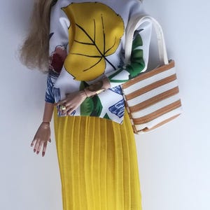 May include: A doll wearing a white and yellow floral print top, a yellow pleated skirt, black sunglasses, and brown earrings. The doll is holding a striped tote bag.