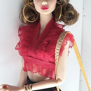Op de afbeelding: Een modepop met lang bruin haar, gekleed in een rood-zwarte kanten jurk, gouden oorringen en een zwarte zonnebril. Ze houdt een bruine leren handtas vast met een gouden gesp.