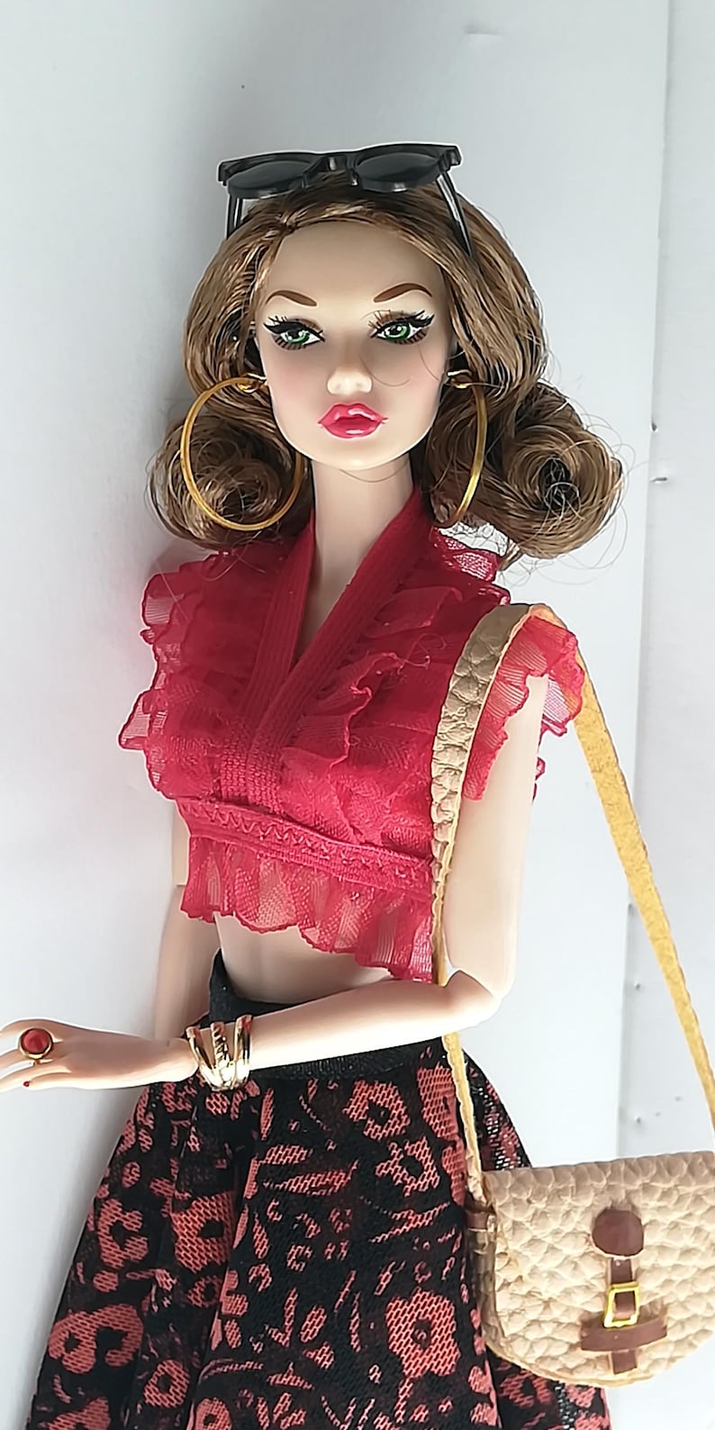 Op de afbeelding: Een modepop met lang bruin haar, gekleed in een rood-zwarte kanten jurk, gouden oorringen en een zwarte zonnebril. Ze houdt een bruine leren handtas vast met een gouden gesp.