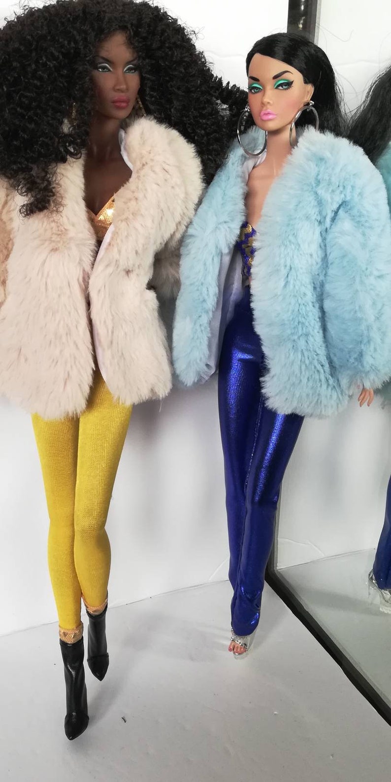 Puede incluir: Dos mu&ntilde;ecas de moda con abrigos de piel sint&eacute;tica. La mu&ntilde;eca de la izquierda lleva un abrigo de color crema y leggings amarillos. La mu&ntilde;eca de la derecha lleva un abrigo azul claro y leggings azules metalizados.