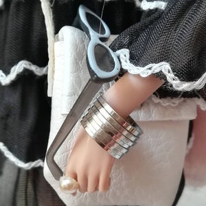 May include: A white faux leather bag with a silver chain strap and a pair of sunglasses tucked into the top. The bag is being held by a person wearing a black dress with white lace trim and a silver crescent moon necklace.