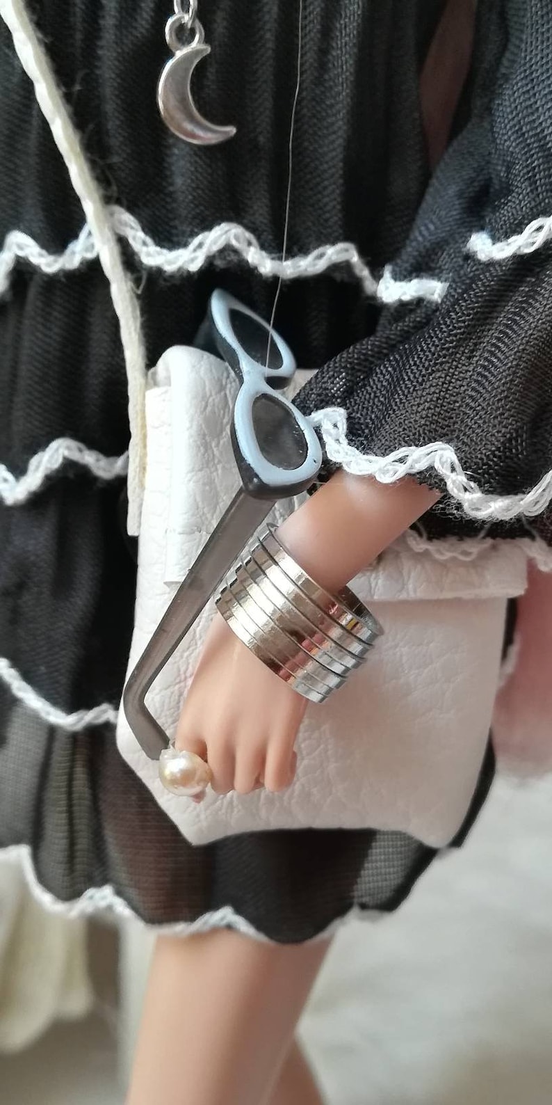 May include: A white faux leather bag with a silver chain strap and a pair of sunglasses tucked into the top. The bag is being held by a person wearing a black dress with white lace trim and a silver crescent moon necklace.
