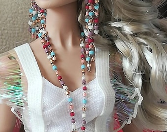 May include: A doll with blonde hair wearing a white dress with a beaded necklace and earrings. The necklace and earrings are multi-colored with red, blue, pink, and gold beads.