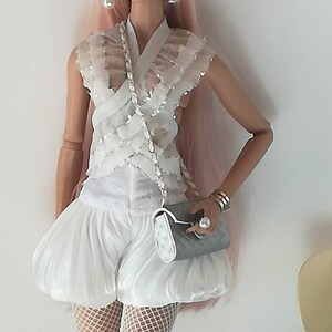 May include: A white doll with pink hair wearing a white ruffled top, white shorts, white fishnet stockings, and white shoes. The doll is holding a white and gray clutch purse and wearing silver sunglasses and earrings.