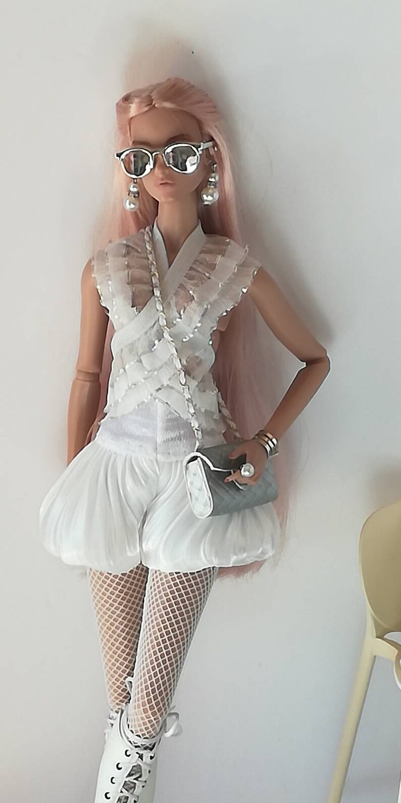 May include: A white doll with pink hair wearing a white ruffled top, white shorts, white fishnet stockings, and white shoes. The doll is holding a white and gray clutch purse and wearing silver sunglasses and earrings.