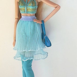 May include: A doll wearing a blue and gold top, a layered blue tulle skirt, blue thigh-high boots, and sunglasses. The doll is holding a blue handbag.