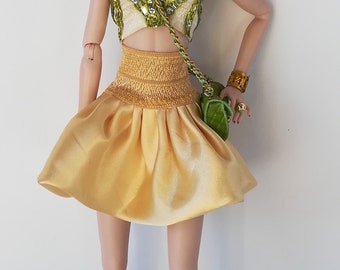 May include: A fashion doll wearing a gold satin skirt, a cream-colored top with green and silver accents, and matching green and gold ankle boots. The doll has brown hair, green eyes, and red lipstick. She is also wearing earrings, a bracelet, and a green purse.