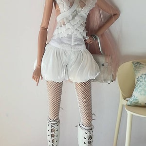 May include: A fashion doll wearing a white lace top, white shorts, white fishnet stockings, and white lace-up boots. The doll has pink hair and is holding a white handbag.
