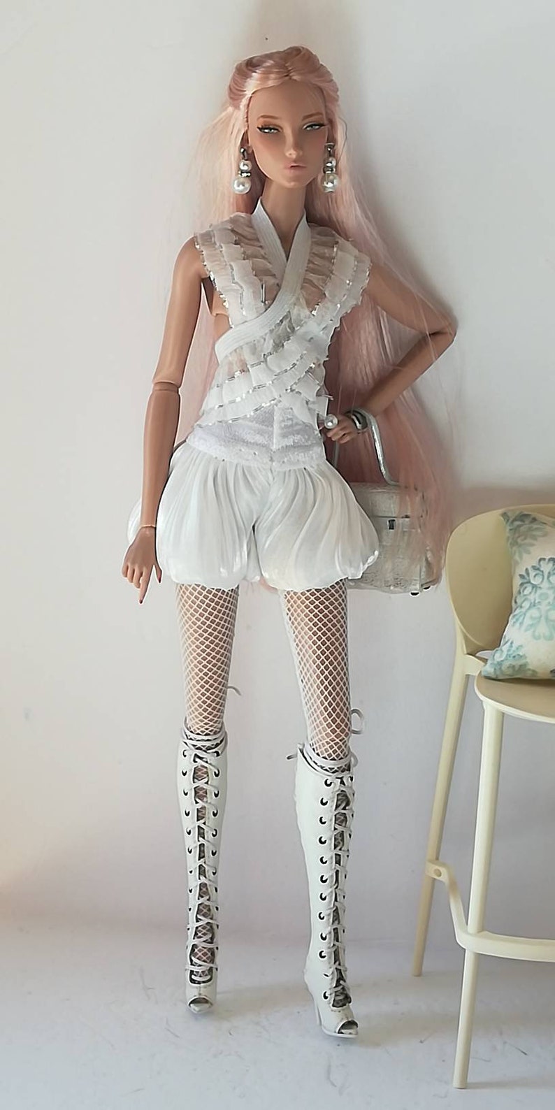 May include: A fashion doll wearing a white lace top, white shorts, white fishnet stockings, and white lace-up boots. The doll has pink hair and is holding a white handbag.
