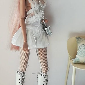 May include: A fashion doll with pink hair wearing a white lace dress, white fishnet tights, and white lace-up boots. The doll is holding a silver handbag and wearing sunglasses.