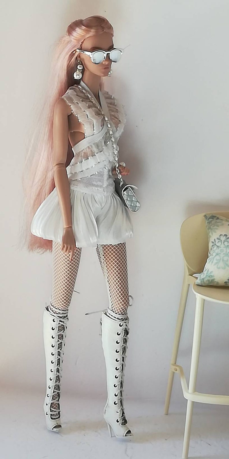 May include: A fashion doll with pink hair wearing a white lace dress, white fishnet tights, and white lace-up boots. The doll is holding a silver handbag and wearing sunglasses.
