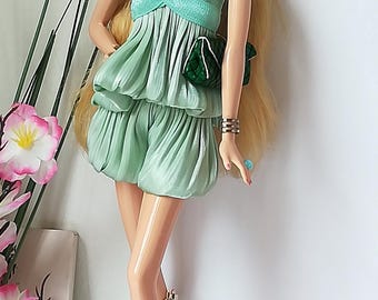 May include: A fashion doll with long blonde hair, wearing a mint green off-the-shoulder top and matching shorts. The doll is accessorized with a green necklace, earrings, bracelet, ring, and a small green purse. She is wearing floral print boots.