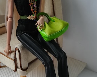May include: A fashion doll wearing a black cap, black top, black pants, and green and yellow boots. The doll is holding a green handbag and is sitting on a white chair.