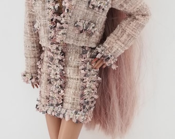 May include: A fashion doll wearing a light pink tweed jacket and matching skirt. The jacket has gold trim and the skirt has a ruffled hem. The doll is wearing brown knee-high boots and pearl earrings. The doll has long pink hair.