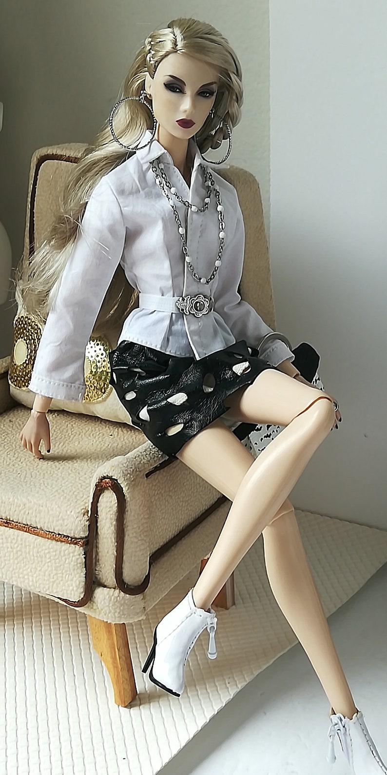 May include: A fashion doll wearing a white button-down shirt with a silver chain necklace, a black skirt with white polka dots, and white heeled boots.