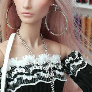 May include: A doll with long pink hair wearing a black and white off-the-shoulder top with a silver chain necklace and a crescent moon pendant. The doll is also wearing large silver hoop earrings.