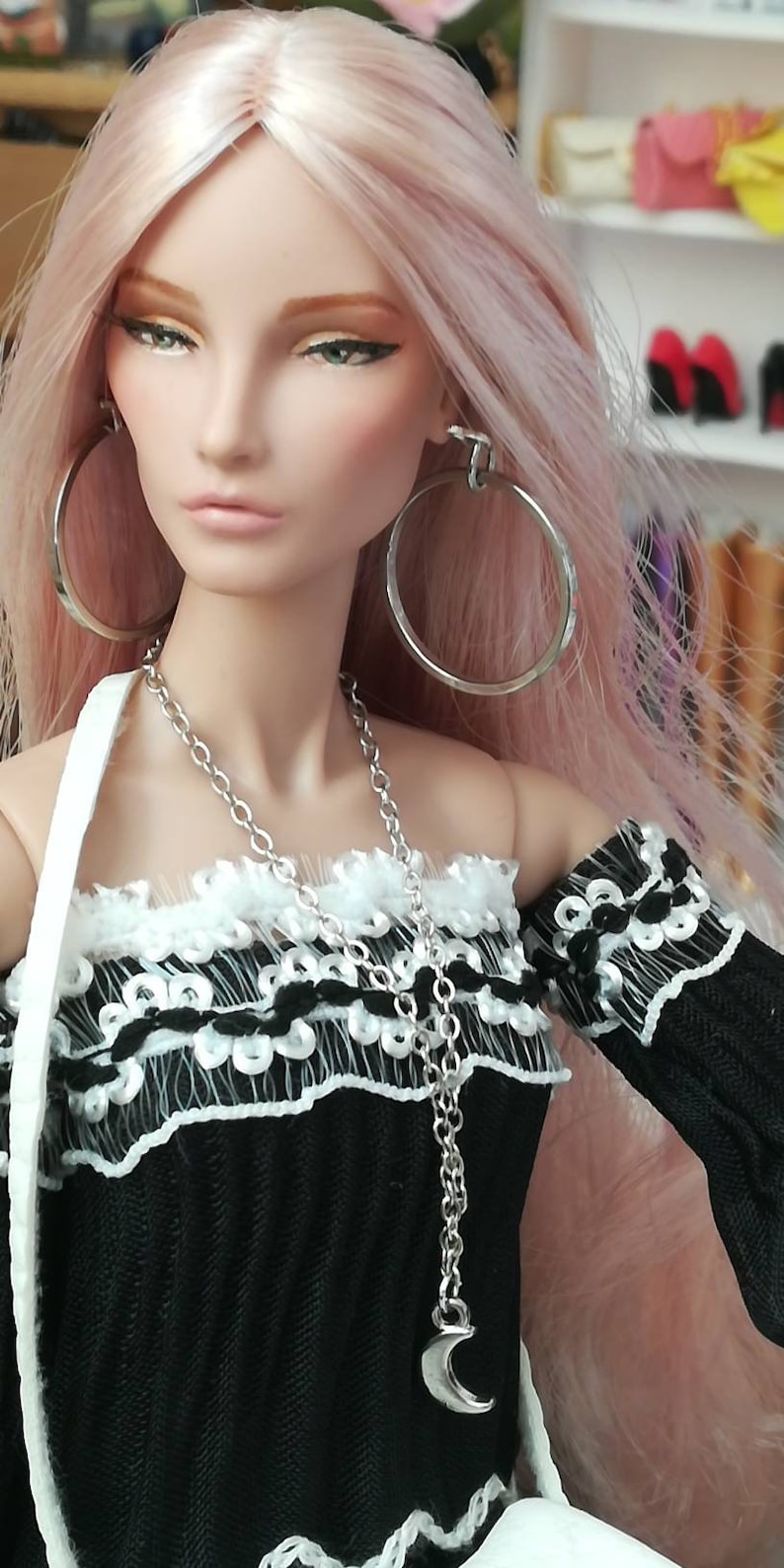 May include: A doll with long pink hair wearing a black and white off-the-shoulder top with a silver chain necklace and a crescent moon pendant. The doll is also wearing large silver hoop earrings.