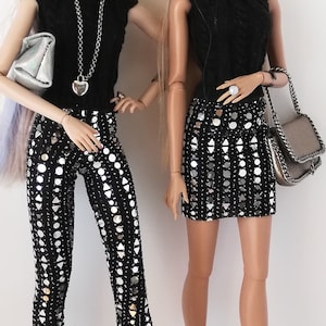 Peut inclure: Deux poupées de mode portant des hauts noirs et des pantalons et une jupe argentés scintillants. La poupée de gauche porte un haut noir avec un collier en chaîne argentée et un pendentif en forme de cœur argenté. La poupée de droite porte un haut noir avec une jupe argentée. Les deux poupées portent des chaussures noires.