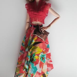 Op de afbeelding: Een modepop met een rood gerimpeld topje, een rok met bloemenprint, gouden oorringen en zwarte sandalen. De pop houdt een bruine clutch vast.