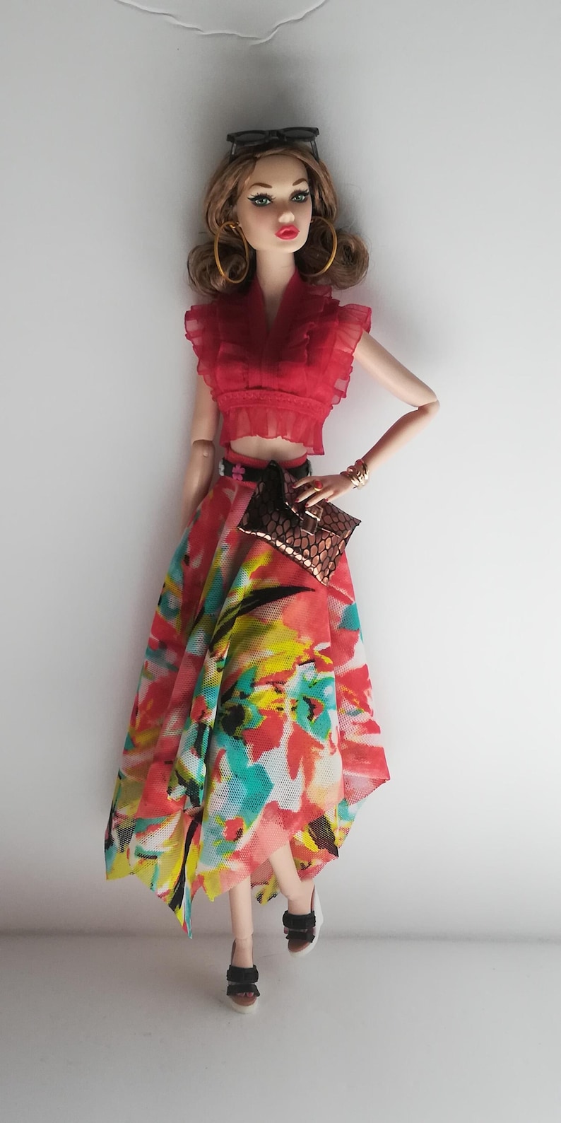 Op de afbeelding: Een modepop met een rood gerimpeld topje, een rok met bloemenprint, gouden oorringen en zwarte sandalen. De pop houdt een bruine clutch vast.