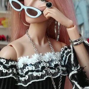 May include: A doll with pink hair wearing a black off-the-shoulder top with white lace trim, a silver necklace with a crescent moon pendant, silver hoop earrings, a black ring with a silver stone, and white sunglasses.