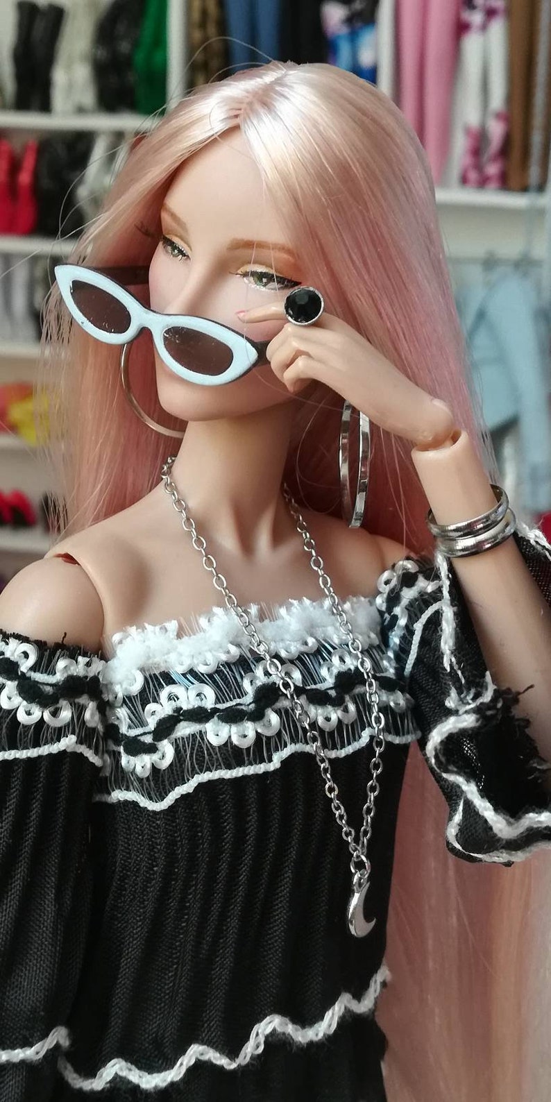 May include: A doll with pink hair wearing a black off-the-shoulder top with white lace trim, a silver necklace with a crescent moon pendant, silver hoop earrings, a black ring with a silver stone, and white sunglasses.