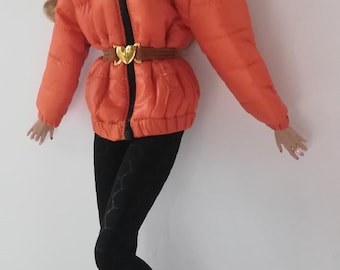 May include: A fashion doll wearing an orange puffer jacket with a black zipper and a brown belt with a gold buckle. The doll is also wearing black leggings, black fringed boots, and orange earmuffs.