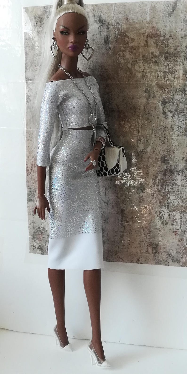 Puede incluir: Un vestido plateado con lentejuelas, falda blanca y bolso blanco. La mu&ntilde;eca lleva pendientes de coraz&oacute;n plateados y un collar de cadena plateada.