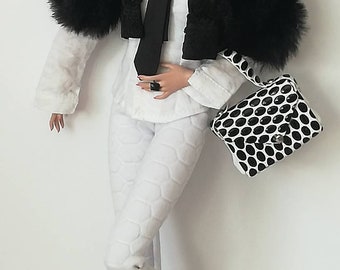 May include: A fashion doll with red hair styled in a ponytail, wearing a black fur jacket, a white shirt with a black tie, white pants, and black boots. The doll is holding a black and white polka dot handbag.