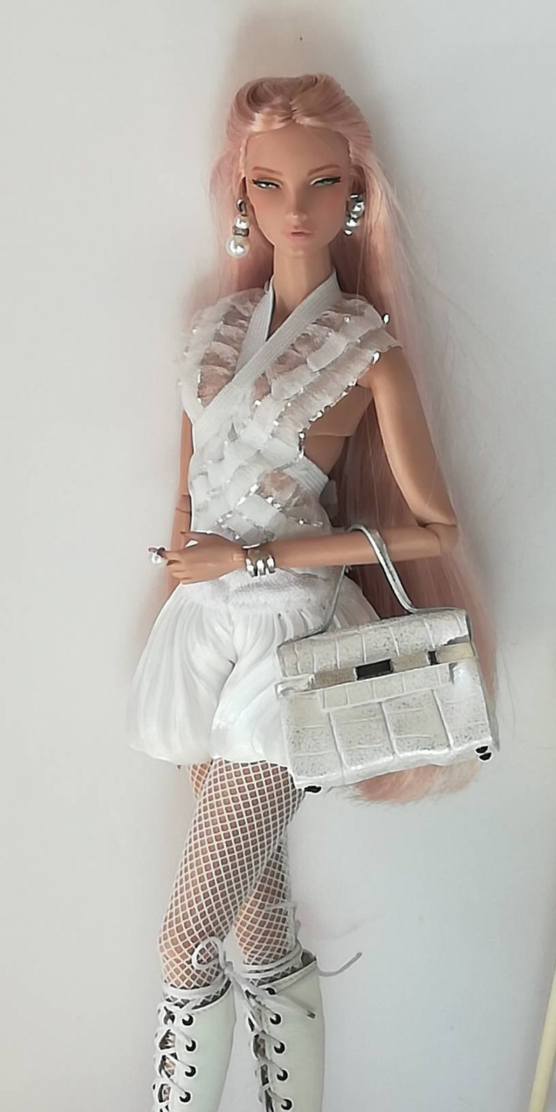 May include: A white doll with pink hair wearing a white lace top, white shorts, white fishnet stockings, and white boots. The doll is holding a white handbag.
