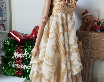 May include: A doll wearing a beige crochet dress with a white and beige patterned overlay, brown hair, and white boots. The doll is wearing a colorful beaded necklace and gold hoop earrings.