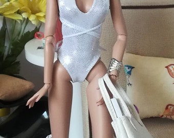 Può includere: Una bambola di moda con i capelli rosa raccolti in una coda di cavallo, indossa un costume da bagno intero bianco scintillante e stivali con tacco trasparente. La bambola tiene una borsa bianca.