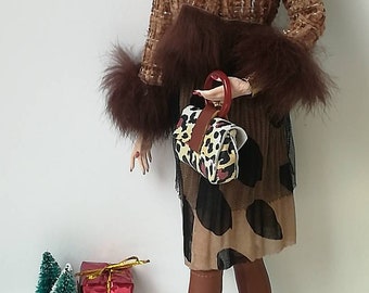 May include: A fashion doll wearing a brown tweed jacket with fur trim, a brown and black leopard print skirt, brown boots, and a leopard print handbag. The doll has black hair and is wearing gold earrings.