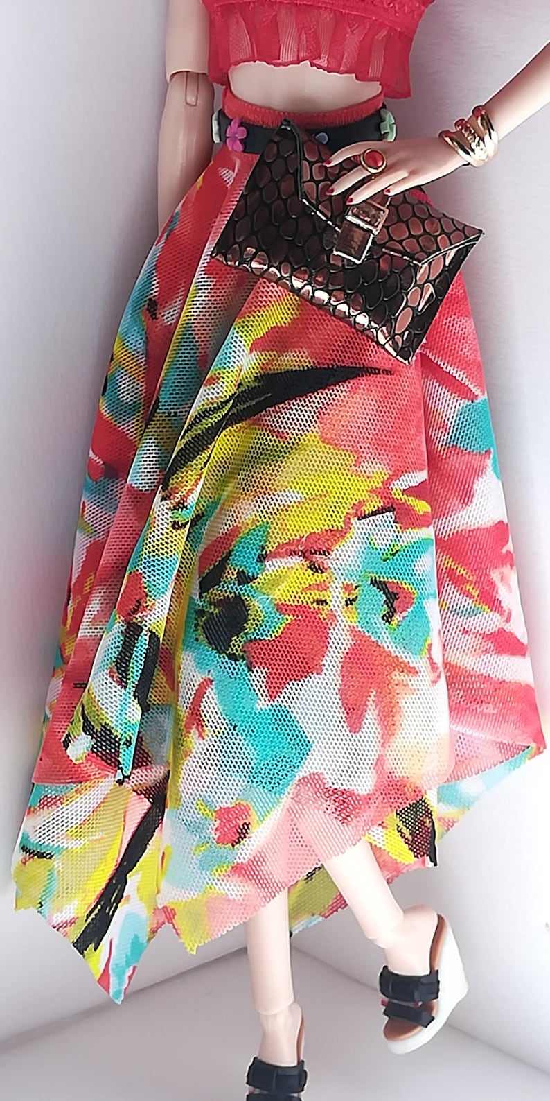 Op de afbeelding: Een pop die een rood topje draagt, een zwarte riem met een bloemenontwerp, een kleurrijke, bloemige mesh-rok en zwarte plateau sandalen. De pop houdt een bruine clutch.