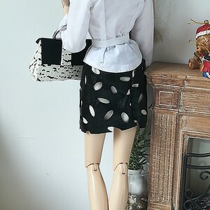 May include: A doll wearing a white button-down shirt, a black skirt with white polka dots, and white heeled boots. The doll is holding a black and white handbag and is standing in front of a miniature fireplace.