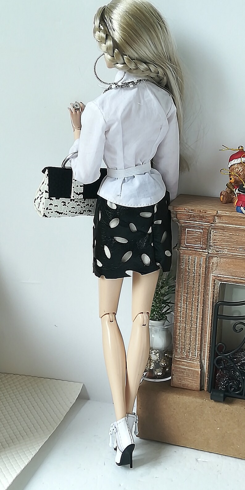 May include: A doll wearing a white button-down shirt, a black skirt with white polka dots, and white heeled boots. The doll is holding a black and white handbag and is standing in front of a miniature fireplace.