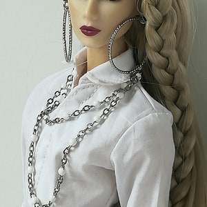 May include: A doll with long blonde hair styled in a braid, wearing a white collared shirt, a silver chain necklace with white beads, a silver belt with a decorative buckle, and large silver hoop earrings.