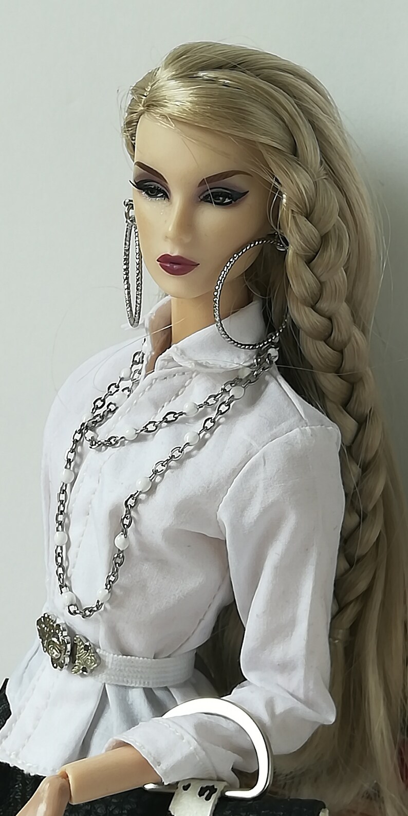 May include: A doll with long blonde hair styled in a braid, wearing a white collared shirt, a silver chain necklace with white beads, a silver belt with a decorative buckle, and large silver hoop earrings.
