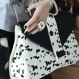 May include: A black and white spotted handbag with a silver clasp and a silver ring with a white stone. The bag is held by a hand wearing a silver ring with a white stone.