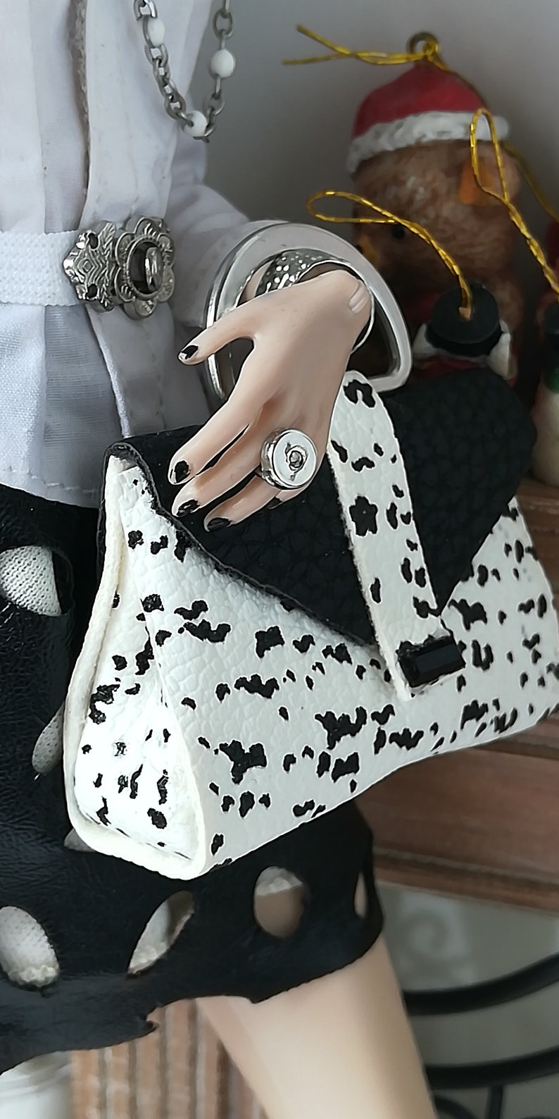 May include: A black and white spotted handbag with a silver clasp and a silver ring with a white stone. The bag is held by a hand wearing a silver ring with a white stone.