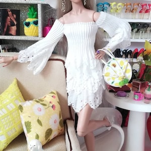 May include: A white doll with blonde hair wearing a white dress with lace trim and a white hat. The doll is holding a yellow and green handbag and is sitting on a white sofa with yellow and white cushions. There is a white dog figurine sitting on the sofa.