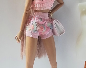 May include: A pink and white doll wearing a pink ruffled top and matching shorts with floral accents. The doll has long pink hair and is wearing pink earrings. The doll is holding a white purse and is wearing pink wedge sandals.
